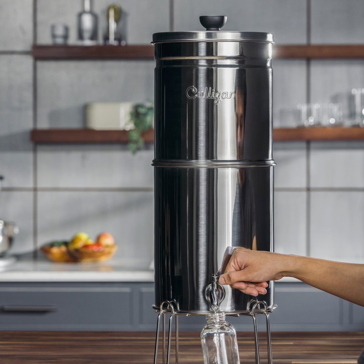 Stainless steel gravity water filter on a kitchen counter with a hand holding a clear container.