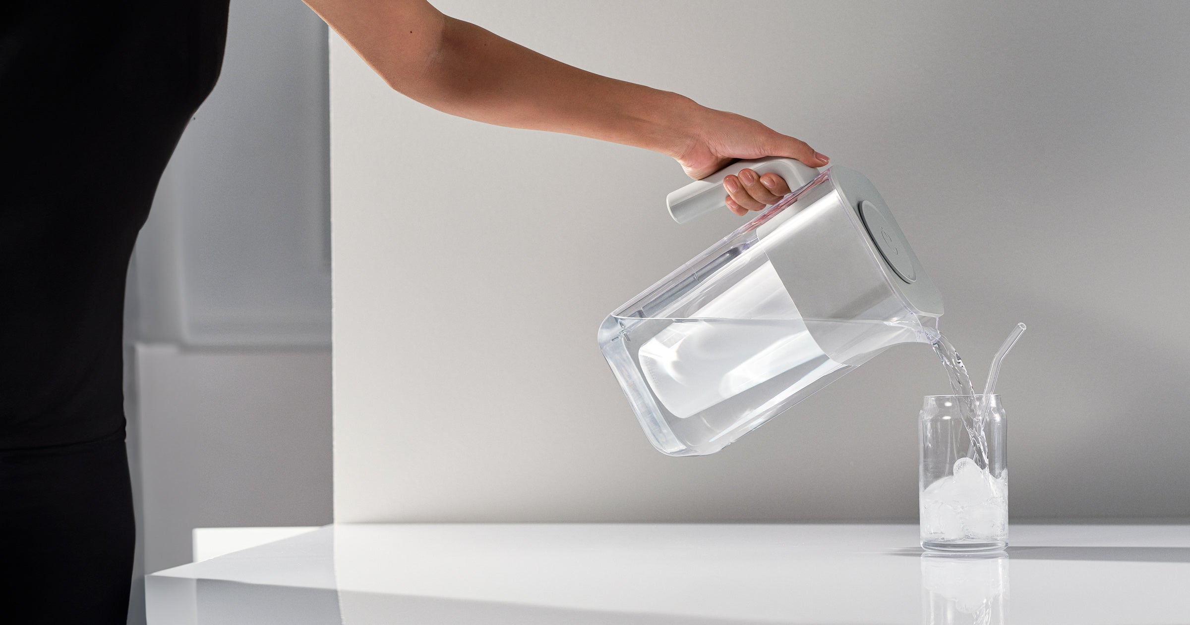 Person pouring water from a clear glass pitcher into a glass on a white surface.